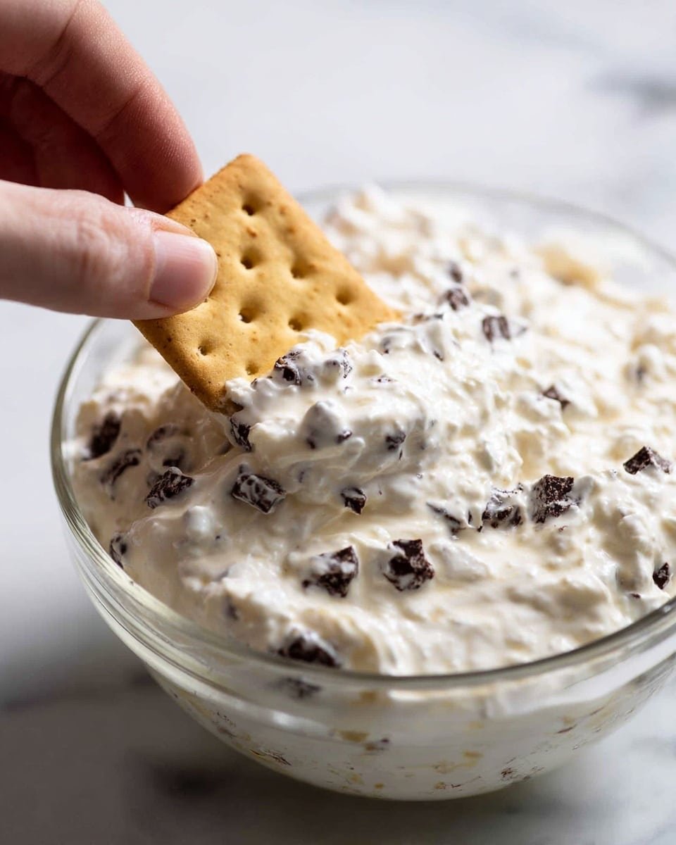 A close-up image shows a clear glass bowl filled with thick, white creamy dip mixed with small dark chocolate chips. A rectangular, light brown cracker with small holes is dipped halfway into the creamy mixture. A woman's hand holds the cracker, positioned on the left side of the image. The background is a white marbled surface. photo taken with an iphone --ar 4:5 --v 7