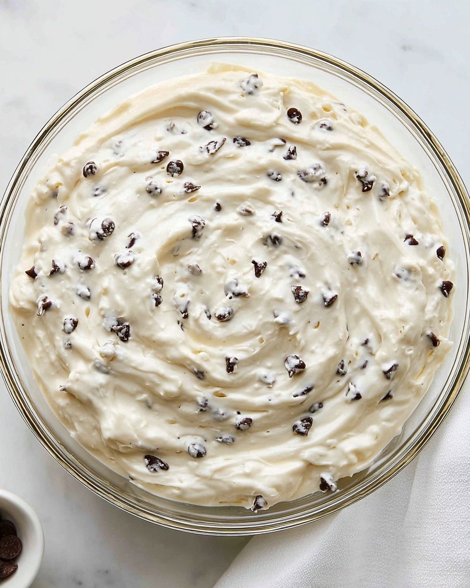 A clear glass bowl filled with a creamy white mixture that is thick and smooth with small dark chocolate chips spread evenly throughout. The texture looks soft and slightly whipped, with gentle swirls on the surface showing the creaminess. The bowl sits on a white marbled surface next to a white cloth napkin partially visible on the right side. Photo taken with an iphone --ar 4:5 --v 7